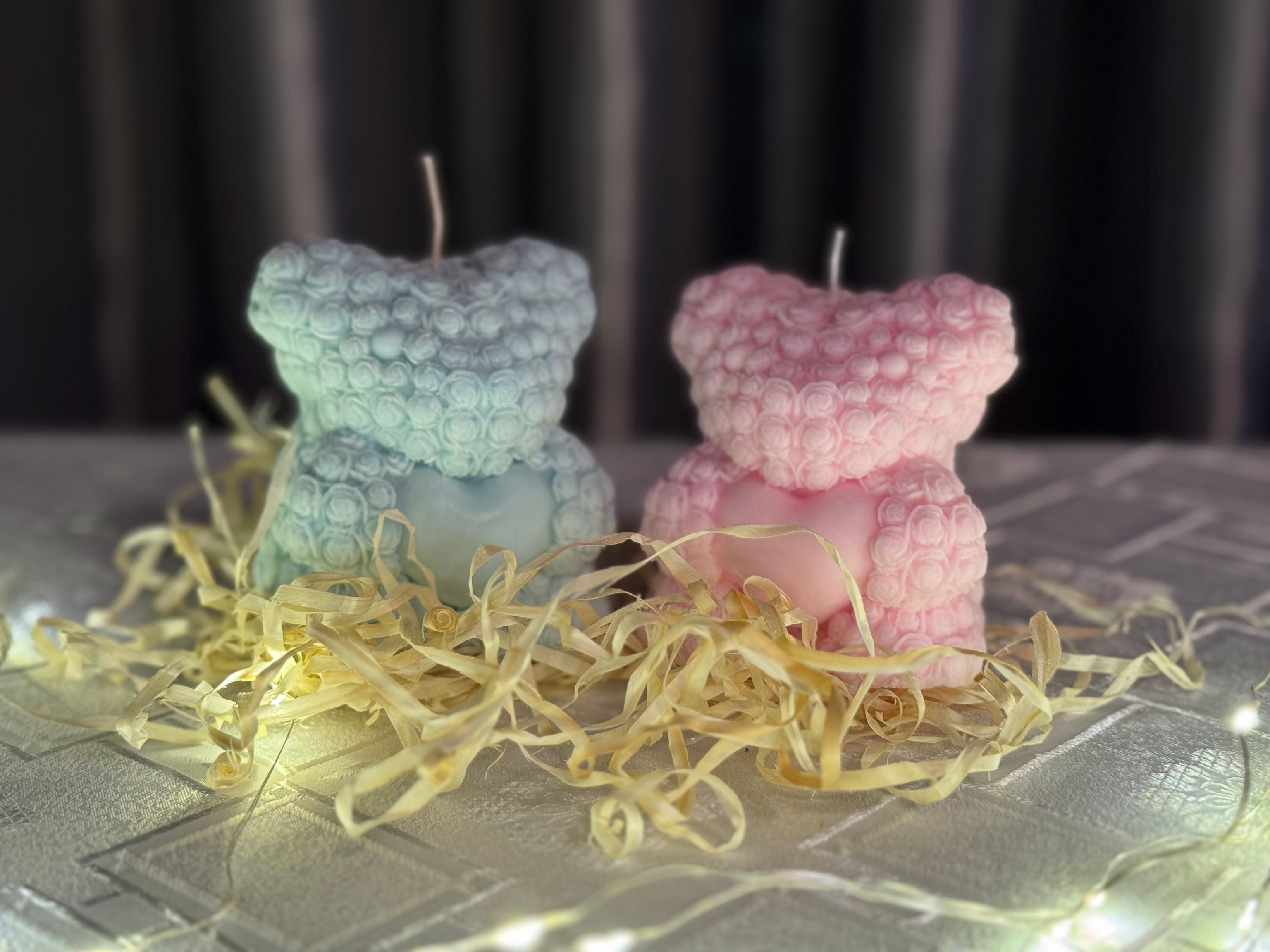 Dwie świece sojowe w kształcie misia stojące na stoliku. Jeden jest błękitny, a drugi różowy. Świece stoją wśród wełny drzewnej.
