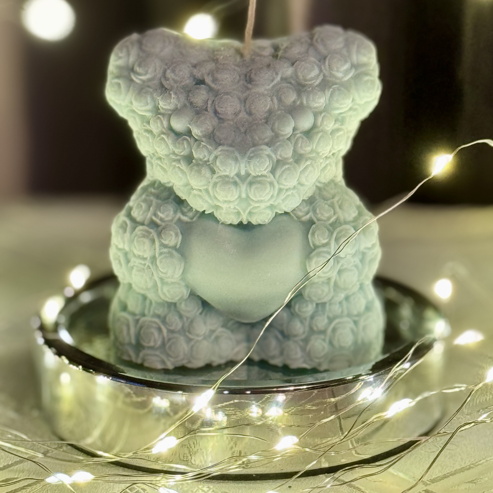 Wolnostojąca świeca sojowa w kształcie misia w błękitnym kolorze stojąca na srebrnej podstawce. Wokół widać błyszczące światełka.