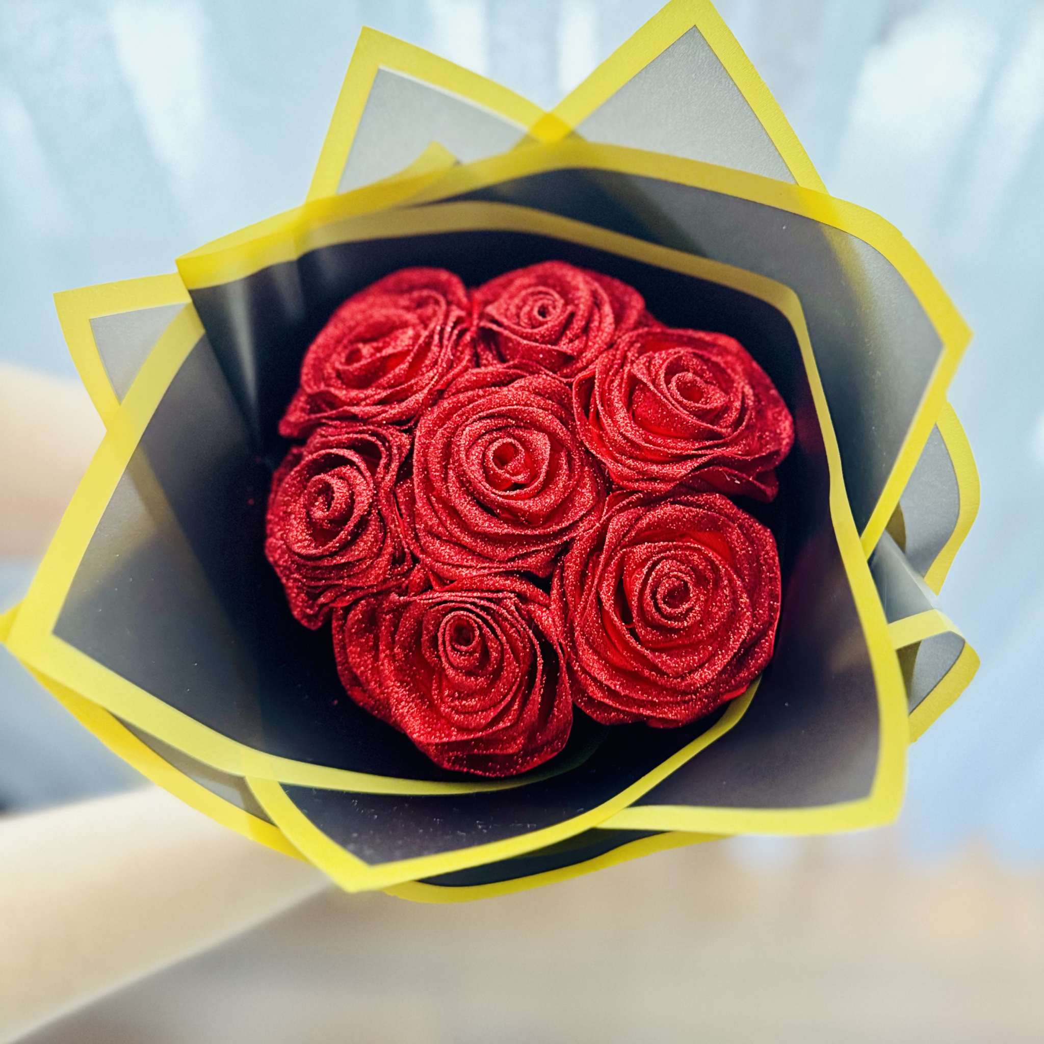 Osoba na zdjęciu trzyma nieduży lecz wspaniale błyszczący bukiet z siedmiu róż, które mienią się dzięki brokatowi, którym są pokryte.