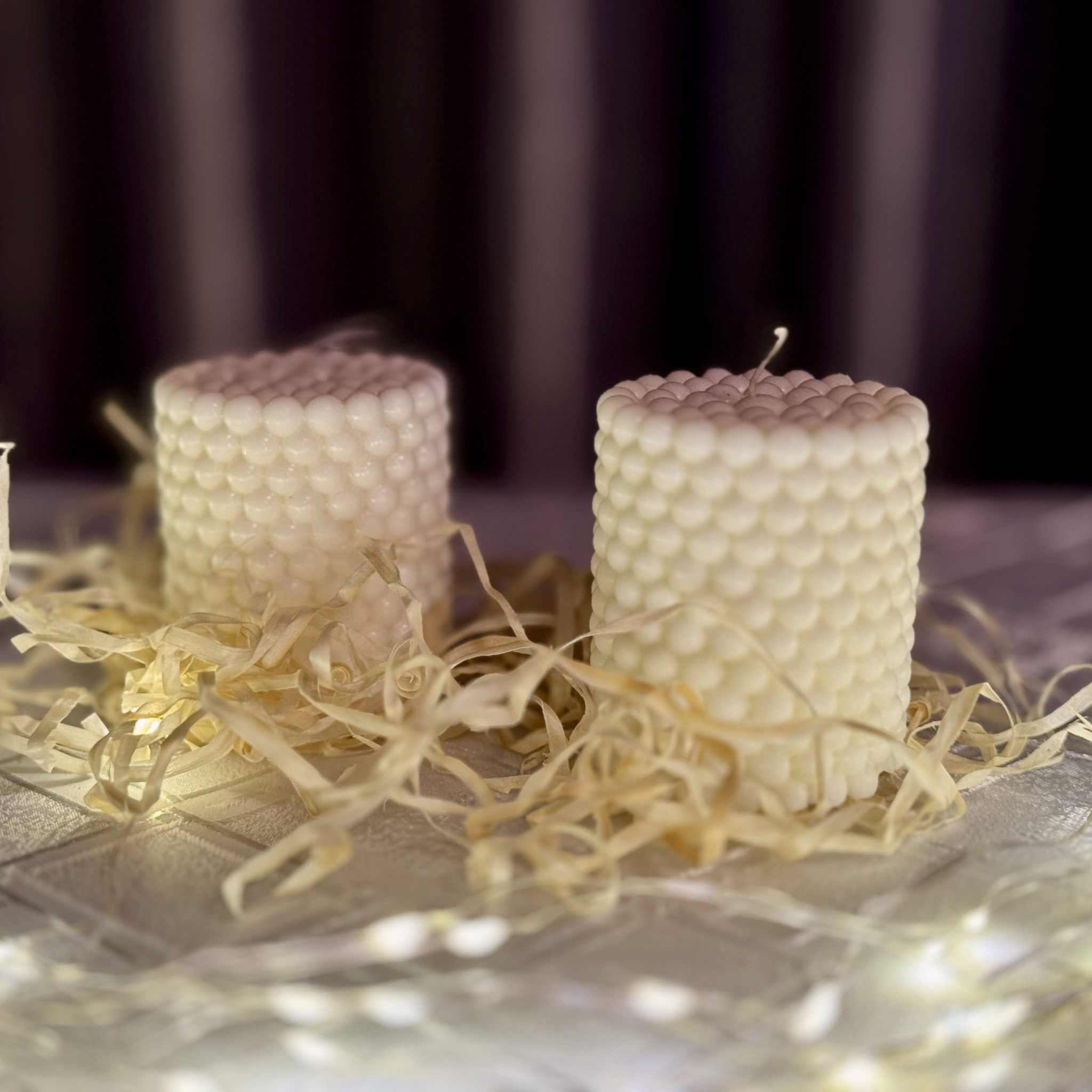 Dwie świece sojowe w naturalnym kolorze wosku w kształcie bąbelkowego walca stojące na stoliku wśród wełny drzewnej.